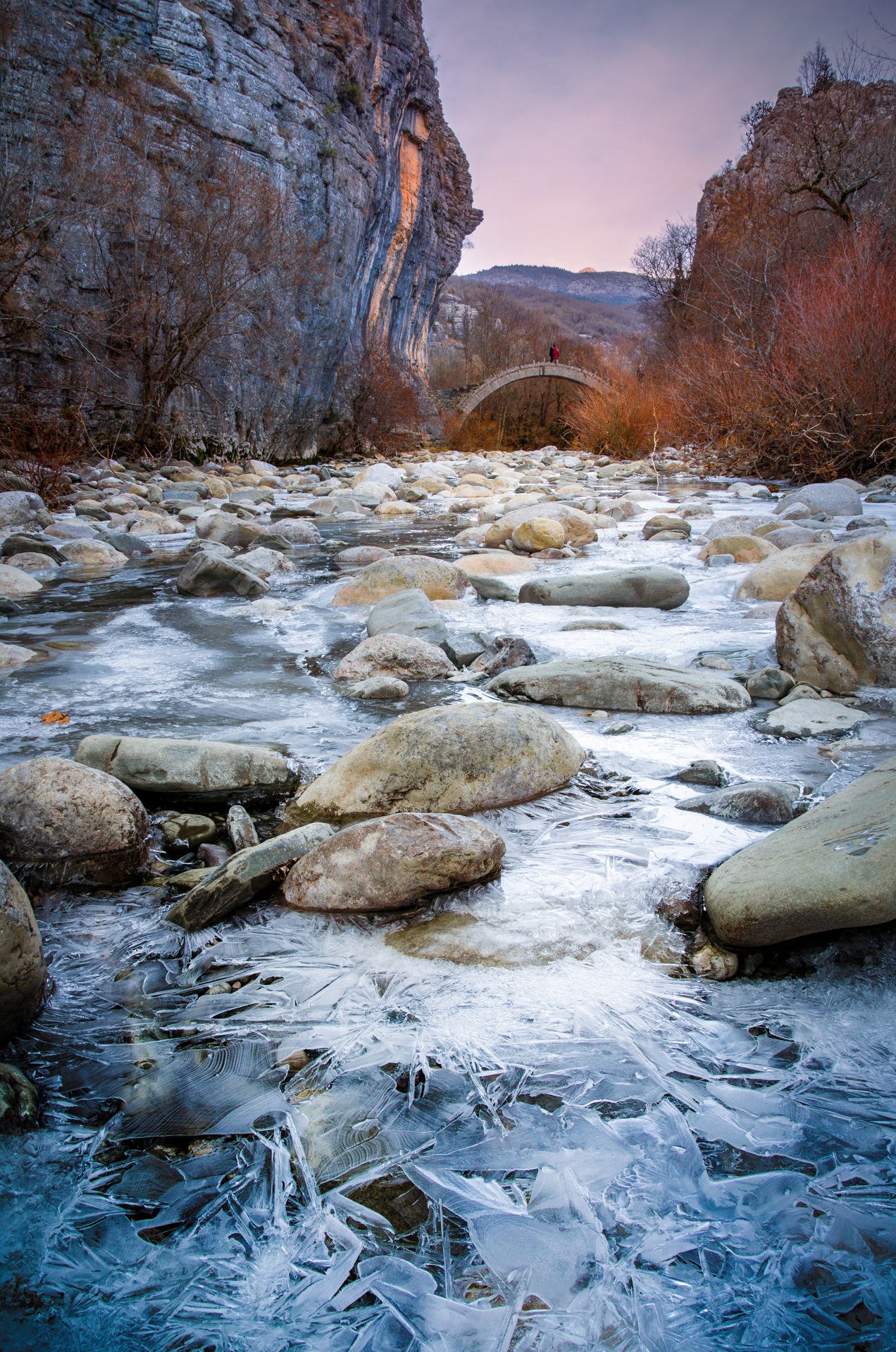 Old Lazaridi - Kontodimou arched stone bridge on Vikos canyon, Zagorohoria, Greece. Winter sunset with ice and glacier on the river