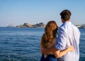 A couple stands arm in arm, facing the sea and gazing at the Old Fortress of Corfu, Greece, with a tall sailing ship anchored nearby under a clear blue sky.