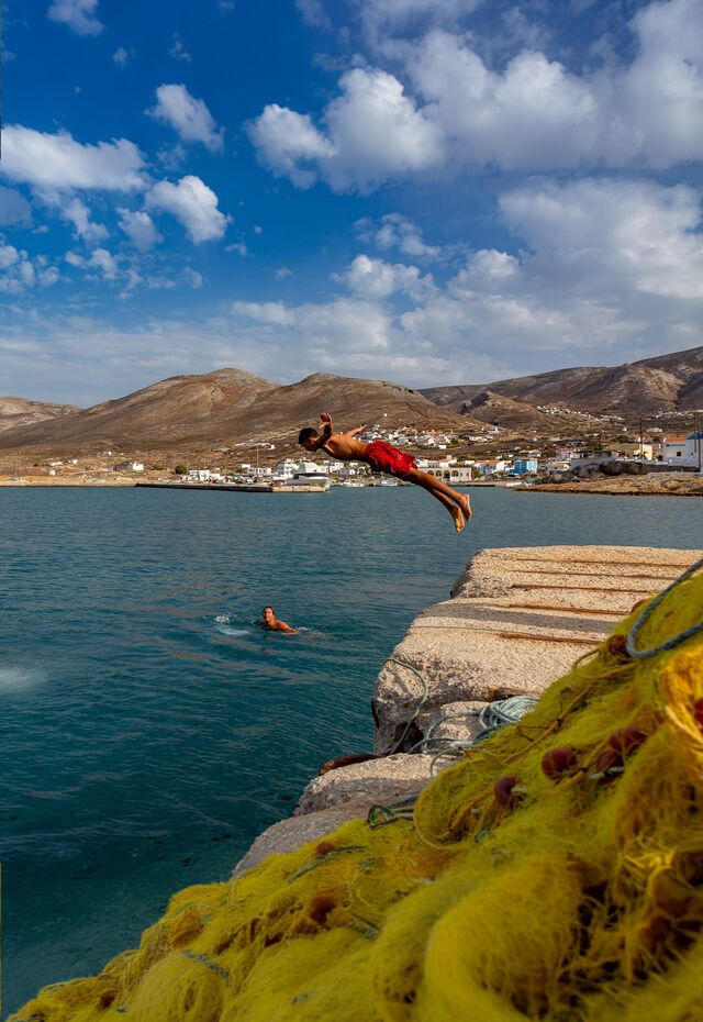 Boy diving off a pier into the sea in Kasos, with mountains and a village in the background.