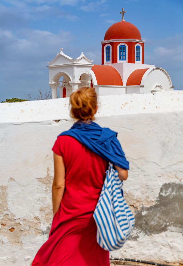 A woman in a red dress with a blue shawl admires a church with red domes and blue windows on Kasos Island.