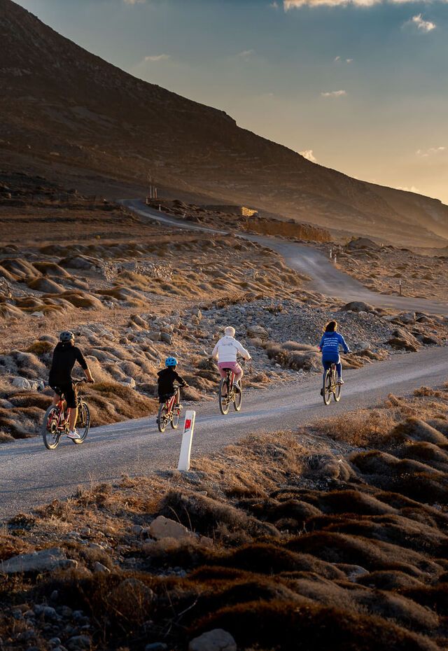 A family rides bicycles along a winding coastal road on Kasos Island, Greece, at sunset, with rugged terrain and the sea in the background.