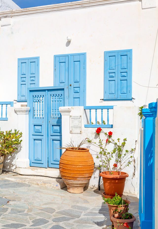 A charming traditional house on Kasos Island, featuring blue shutters and door, surrounded by potted plants.