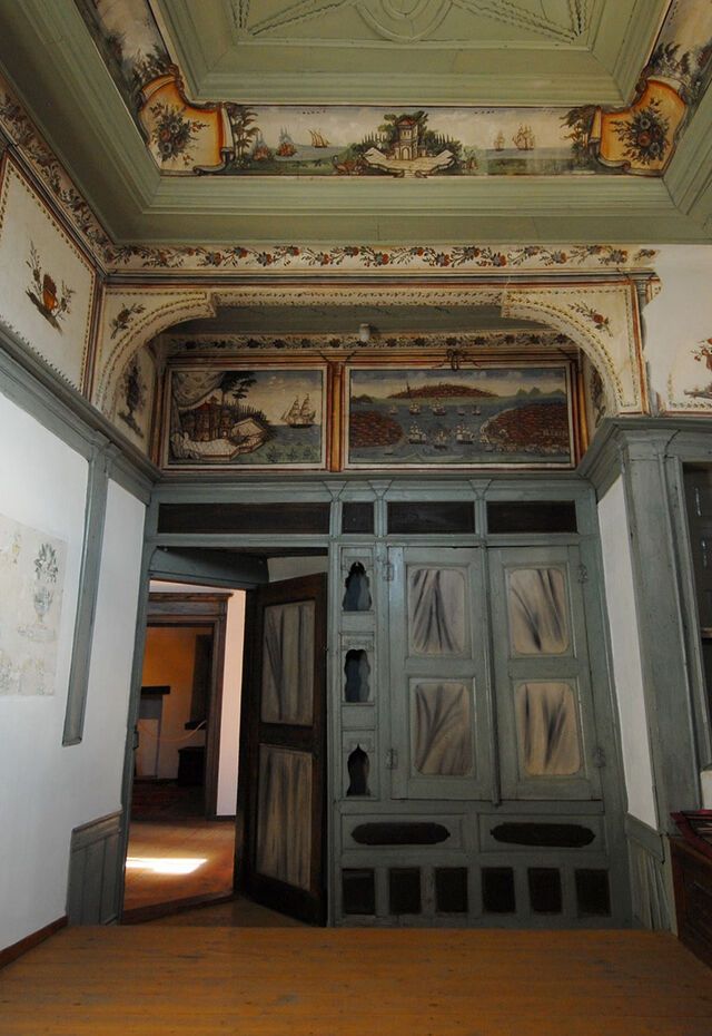 Vareltzidaina Mansion interior showing an ornate painted ceiling frieze, carved doorway and a bench along the wall.
