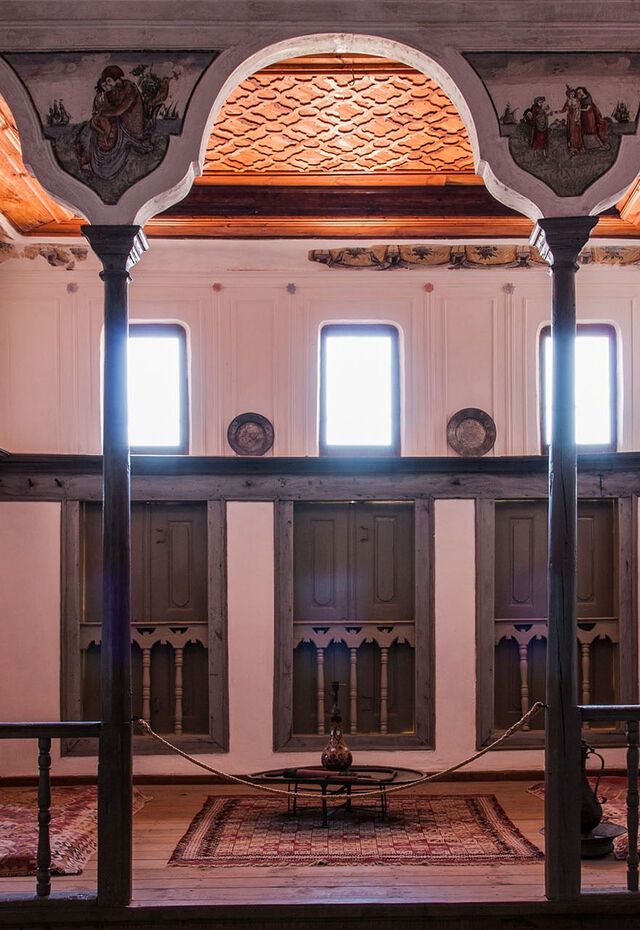 Main hall of Vareltzidaina Mansion with wooden gallery, central columns, arched painted ceiling panels and patterned rugs.